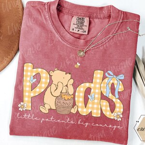 May include: A maroon Comfort Colors t-shirt featuring a graphic of Winnie the Pooh with the word "Pods" in yellow plaid letters. The shirt includes the text "little patients, big courage" and a small bee illustration.