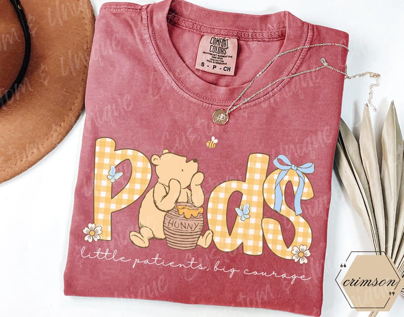 May include: A maroon Comfort Colors t-shirt featuring a graphic of Winnie the Pooh with the word "Pods" in yellow plaid letters. The shirt includes the text "little patients, big courage" and a small bee illustration.