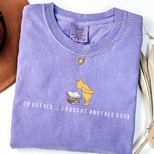 May include: A lilac-colored t-shirt with the phrase "OH BOTHER... I BOUGHT ANOTHER BOOK" printed in white. The shirt features a cartoon illustration of Winnie the Pooh looking at a stack of books. A gold necklace and a brown hat are also visible.