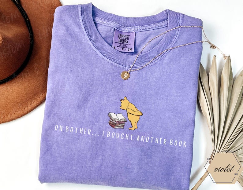 May include: A lilac-colored t-shirt with the phrase "OH BOTHER... I BOUGHT ANOTHER BOOK" printed in white. The shirt features a cartoon illustration of Winnie the Pooh looking at a stack of books. A gold necklace and a brown hat are also visible.