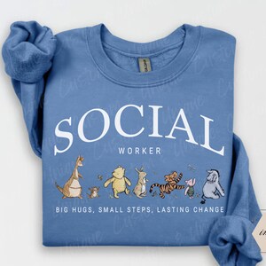 Pu&ograve; includere: Una felpa blu chiaro con la scritta bianca "SOCIAL WORKER". Sotto, personaggi dei cartoni animati. La frase "BIG HUGS, SMALL STEPS, LASTING CHANGE" &egrave; stampata in basso. La felpa ha le maniche lunghe.