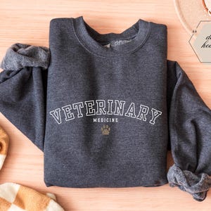 May include: Dark grey sweatshirt featuring the words "VETERINARY MEDICINE" in white, arched across the chest, with a paw print graphic below. The sleeves are cuffed. The words "dark heather" are in a hexagon-shaped graphic.