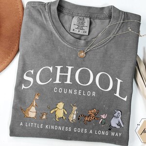 May include: A charcoal gray t-shirt featuring the white text SCHOOL COUNSELOR and the phrase A LITTLE KINDNESS GOES A LONG WAY. Below the text are illustrations of Kanga, Roo, Winnie the Pooh, Rabbit, Tigger, Piglet, and Eeyore in muted colors.