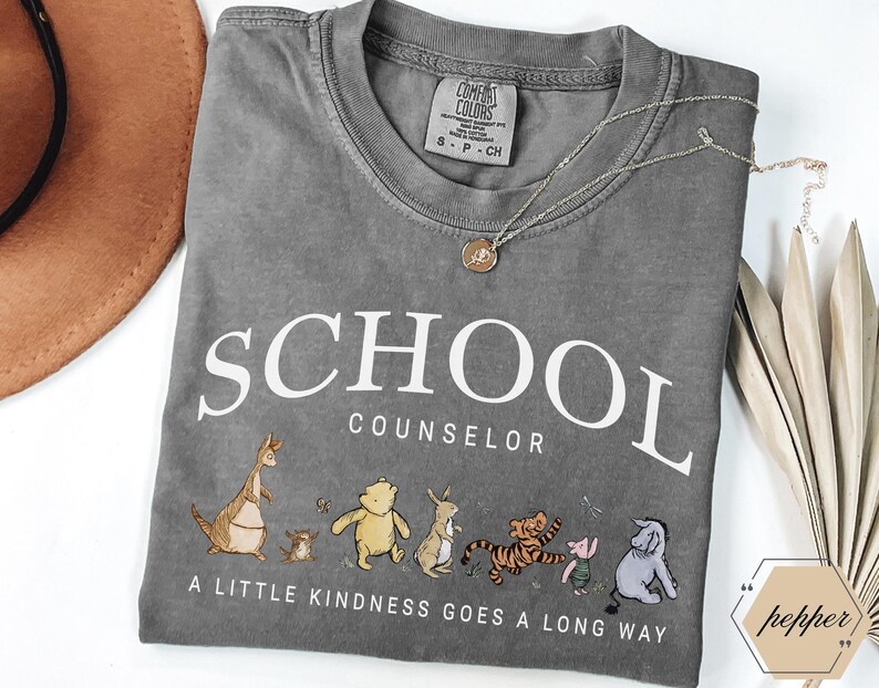 May include: A charcoal gray t-shirt featuring the white text SCHOOL COUNSELOR and the phrase A LITTLE KINDNESS GOES A LONG WAY. Below the text are illustrations of Kanga, Roo, Winnie the Pooh, Rabbit, Tigger, Piglet, and Eeyore in muted colors.