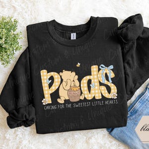 May include: Black sweatshirt with the word "Pads" in yellow gingham print, featuring a cartoon bear with a honey pot. The text "Caring for the sweetest little hearts" is below the word "Pads". A small bee and blue bow are also visible.