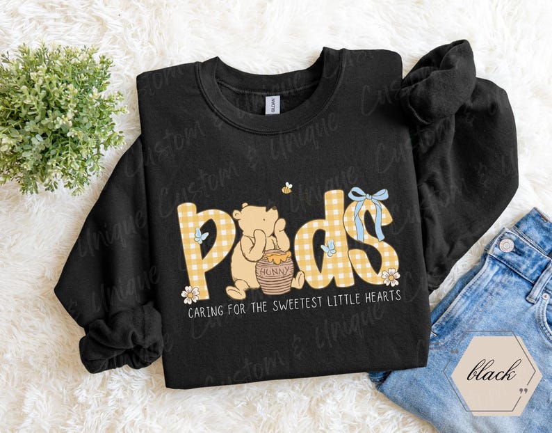 May include: Black sweatshirt with the word "Pads" in yellow gingham print, featuring a cartoon bear with a honey pot. The text "Caring for the sweetest little hearts" is below the word "Pads". A small bee and blue bow are also visible.
