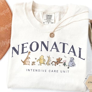 May include: An ivory t-shirt featuring the dark blue text NEONATAL above a graphic of Winnie the Pooh characters including Roo, Piglet, Pooh, Rabbit, Tigger, and Eeyore, with the text INTENSIVE CARE UNIT printed below the characters.