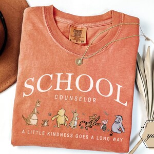 May include: A terracotta-colored t-shirt featuring the white text SCHOOL COUNSELOR and a graphic of Winnie the Pooh characters. Below the characters, the text reads A LITTLE KINDNESS GOES A LONG WAY. The shirt has a Comfort Colors brand label.