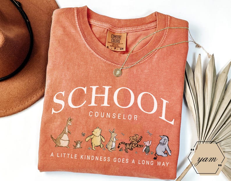 May include: A terracotta-colored t-shirt featuring the white text SCHOOL COUNSELOR and a graphic of Winnie the Pooh characters. Below the characters, the text reads A LITTLE KINDNESS GOES A LONG WAY. The shirt has a Comfort Colors brand label.