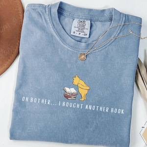 May include: A light blue Comfort Colors t-shirt featuring a graphic of Winnie the Pooh and books, with the text "OH BOTHER... I BOUGHT ANOTHER BOOK." A gold necklace rests on the shirt. The shirt is a size small.