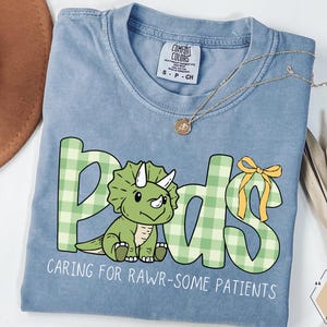 May include: A light blue Comfort Colors t-shirt with a green dinosaur and the word "Pods" in a green plaid pattern. The text "CARING FOR RAWR-SOME PATIENTS" is below the dinosaur. A gold necklace is draped across the shirt.