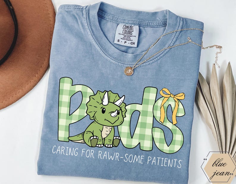 May include: A light blue Comfort Colors t-shirt with a green dinosaur and the word "Pods" in a green plaid pattern. The text "CARING FOR RAWR-SOME PATIENTS" is below the dinosaur. A gold necklace is draped across the shirt.