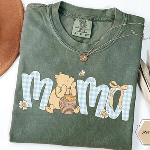May include: A sage green t-shirt with the word "mama" in a playful font, featuring a Winnie the Pooh graphic. The letters have a blue and white gingham pattern. A gold necklace and a straw hat are also visible.