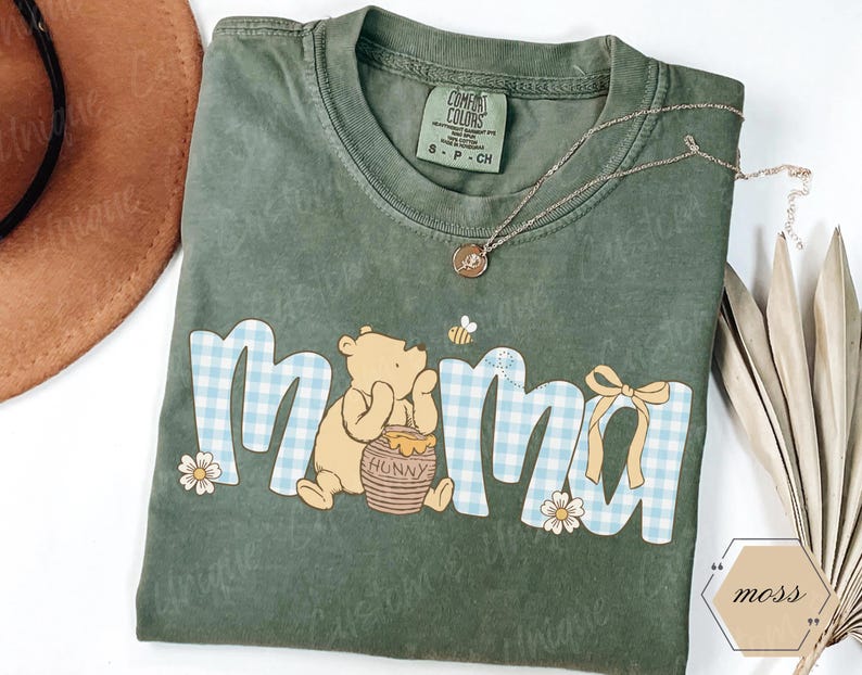 May include: A sage green t-shirt with the word "mama" in a playful font, featuring a Winnie the Pooh graphic. The letters have a blue and white gingham pattern. A gold necklace and a straw hat are also visible.