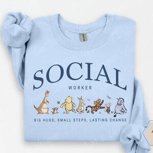 Pu&ograve; includere: Felpa azzurra con le parole "SOCIAL WORKER" in testo blu navy ad arco. Sotto il testo c'&egrave; una fila di personaggi dei cartoni animati. Il testo "BIG HUGS, SMALL STEPS, LASTING CHANGE" &egrave; stampato sotto.