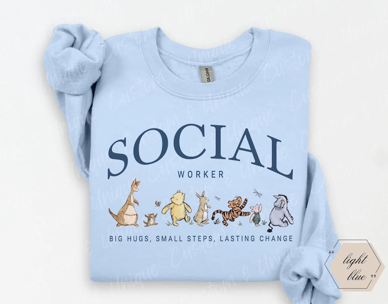 Pu&ograve; includere: Felpa azzurra con le parole "SOCIAL WORKER" in testo blu navy ad arco. Sotto il testo c'&egrave; una fila di personaggi dei cartoni animati. Il testo "BIG HUGS, SMALL STEPS, LASTING CHANGE" &egrave; stampato sotto.
