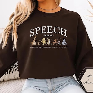May include: Dark chocolate brown sweatshirt with the words "SPEECH THERAPY" in white arched text. Below the text is a row of cartoon characters. The phrase "EVERY WAY TO COMMUNICATE IS THE RIGHT WAY" is printed below the characters.