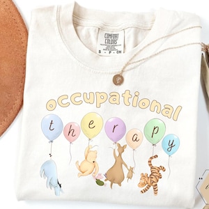 May include: Ivory-colored Comfort Colors t-shirt with the words "occupational therapy" in a playful font. Colorful balloons with cartoon characters are featured below the text. A gold necklace is draped on the shirt.