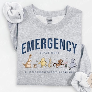 Puede incluir: Una sudadera gris claro con la frase "EMERGENCY DEPARTMENT" en azul marino en la parte superior. Debajo del texto hay personajes de dibujos animados. La frase "A LITTLE KINDNESS GOES A LONG WAY" está impresa debajo de los personajes.