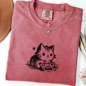 May include: Crimson-colored t-shirt featuring a black graphic of a cat playing a video game. The shirt has a crew neck and short sleeves. A delicate silver necklace with a small pendant rests on the shirt. The shirt is labeled "Comfort Colors".