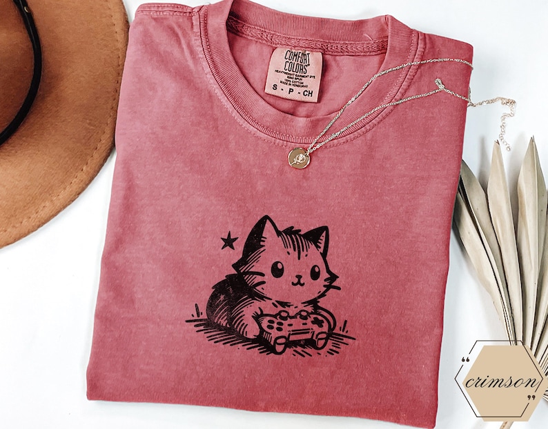 May include: Crimson-colored t-shirt featuring a black graphic of a cat playing a video game. The shirt has a crew neck and short sleeves. A delicate silver necklace with a small pendant rests on the shirt. The shirt is labeled "Comfort Colors".