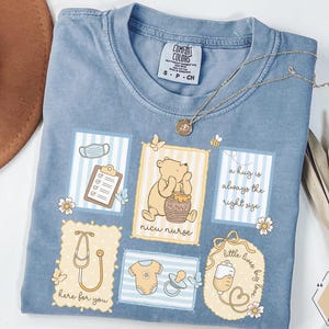 Peut inclure: T-shirt bleu clair Comfort Colors avec des illustrations de Winnie l'Ourson en tant qu'infirmière de soins intensifs néonatals. Le t-shirt présente un presse-papiers, un stéthoscope et le texte "a hug is always the right size". Un collier doré est drapé sur le t-shirt.