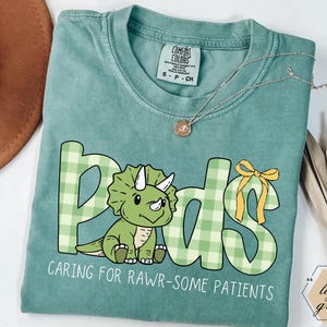May include: A light green t-shirt with the word "Peds" in a playful font, featuring a green dinosaur illustration and the phrase "Caring for Rawr-some Patients." The letters are in a green plaid pattern. A gold necklace is draped across the shirt.
