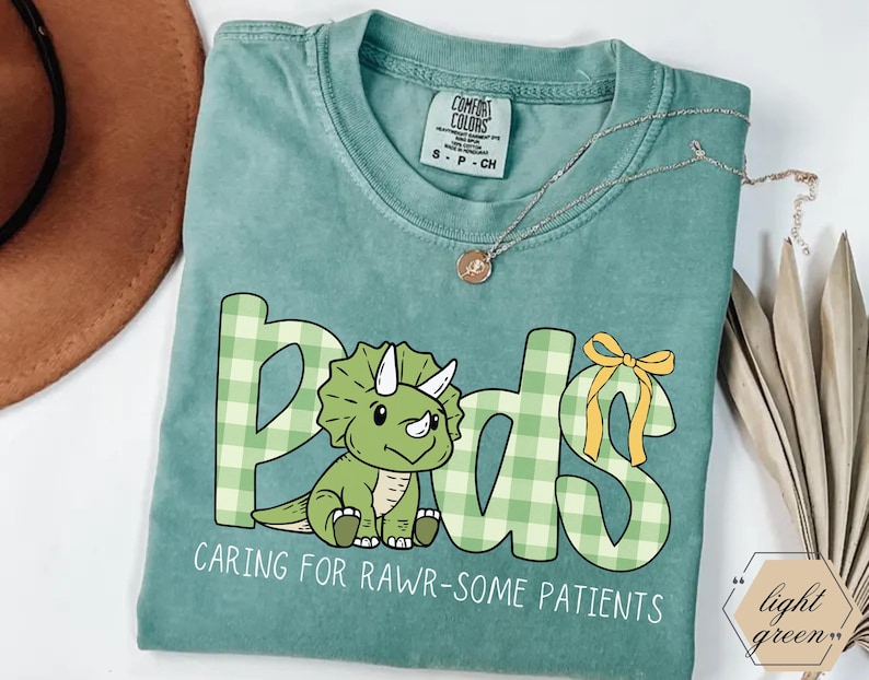 May include: A light green t-shirt with the word "Peds" in a playful font, featuring a green dinosaur illustration and the phrase "Caring for Rawr-some Patients." The letters are in a green plaid pattern. A gold necklace is draped across the shirt.