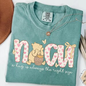 May include: A light green t-shirt with the word "N&CU" in a pink heart pattern, featuring a cartoon bear and the text "a hug is always the right size." A gold necklace rests on the shirt.