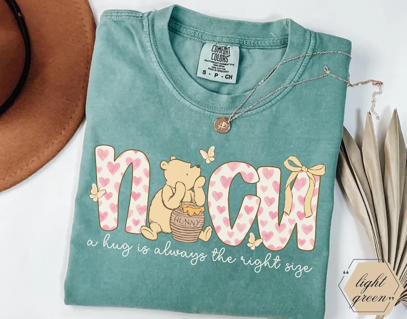 May include: A light green t-shirt with the word "N&CU" in a pink heart pattern, featuring a cartoon bear and the text "a hug is always the right size." A gold necklace rests on the shirt.