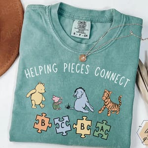 Op de afbeelding: Een saliegroen t-shirt met korte mouwen en een afbeelding van Winnie de Poeh, Knorretje, Iejoor en Teigetje boven vier puzzelstukjes met de letters B, C, B en A. De tekst HELPING PIECES CONNECT staat in het wit boven de figuren.