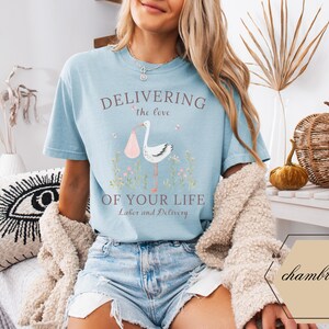 Op de afbeelding: Een lichtblauw t-shirt met de tekst "Delivering the love of your life Labor and Delivery" en een roze en witte ooievaarillustratie.