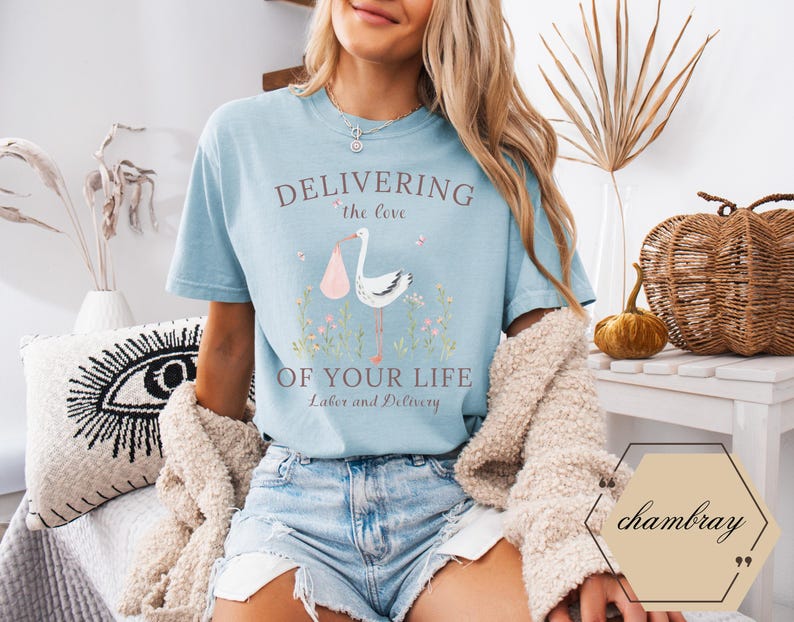 Op de afbeelding: Een lichtblauw t-shirt met de tekst "Delivering the love of your life Labor and Delivery" en een roze en witte ooievaarillustratie.