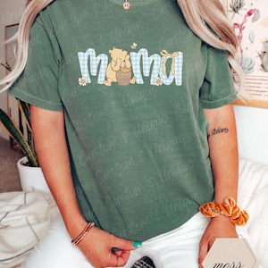 May include: Green short-sleeved t-shirt with the word "mama" in a blue gingham pattern, featuring a cartoon bear holding a honey pot. A small bee and flowers accent the design. The shirt is a comfortable, casual style.