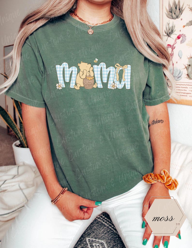 May include: Green short-sleeved t-shirt with the word "mama" in a blue gingham pattern, featuring a cartoon bear holding a honey pot. A small bee and flowers accent the design. The shirt is a comfortable, casual style.