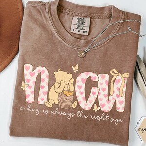 May include: Brown t-shirt with the word "NMCU" in pink and white, decorated with hearts and a cartoon bear. The text "a hug is always the right size" is below the graphic. A silver necklace and a brown hat are also visible.