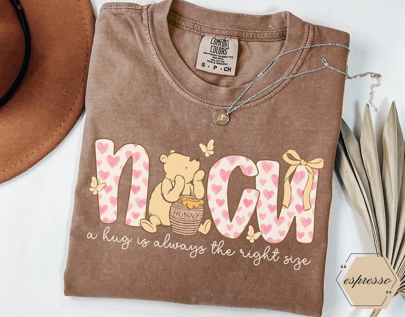 May include: Brown t-shirt with the word "NMCU" in pink and white, decorated with hearts and a cartoon bear. The text "a hug is always the right size" is below the graphic. A silver necklace and a brown hat are also visible.