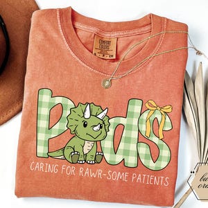 May include: Burnt orange t-shirt with the word "Peds" in green plaid letters, featuring a cartoon triceratops. The text "Caring for Rawr-some Patients" is below the word "Peds". A gold necklace is draped across the shirt.