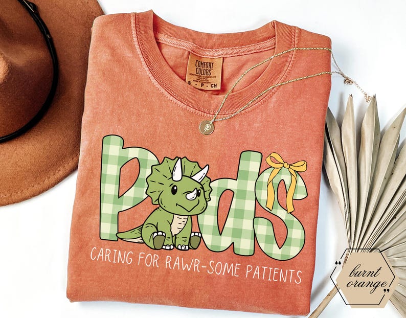 May include: Burnt orange t-shirt with the word "Peds" in green plaid letters, featuring a cartoon triceratops. The text "Caring for Rawr-some Patients" is below the word "Peds". A gold necklace is draped across the shirt.