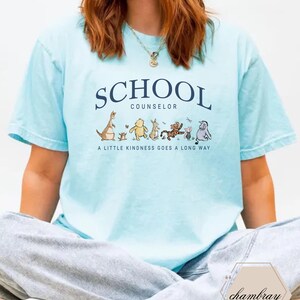 May include: A light blue short-sleeve t-shirt featuring the dark blue text SCHOOL COUNSELOR above a graphic of various cartoon animals and the text A LITTLE KINDNESS GOES A LONG WAY. The shirt has a crew neckline and a relaxed, casual fit.