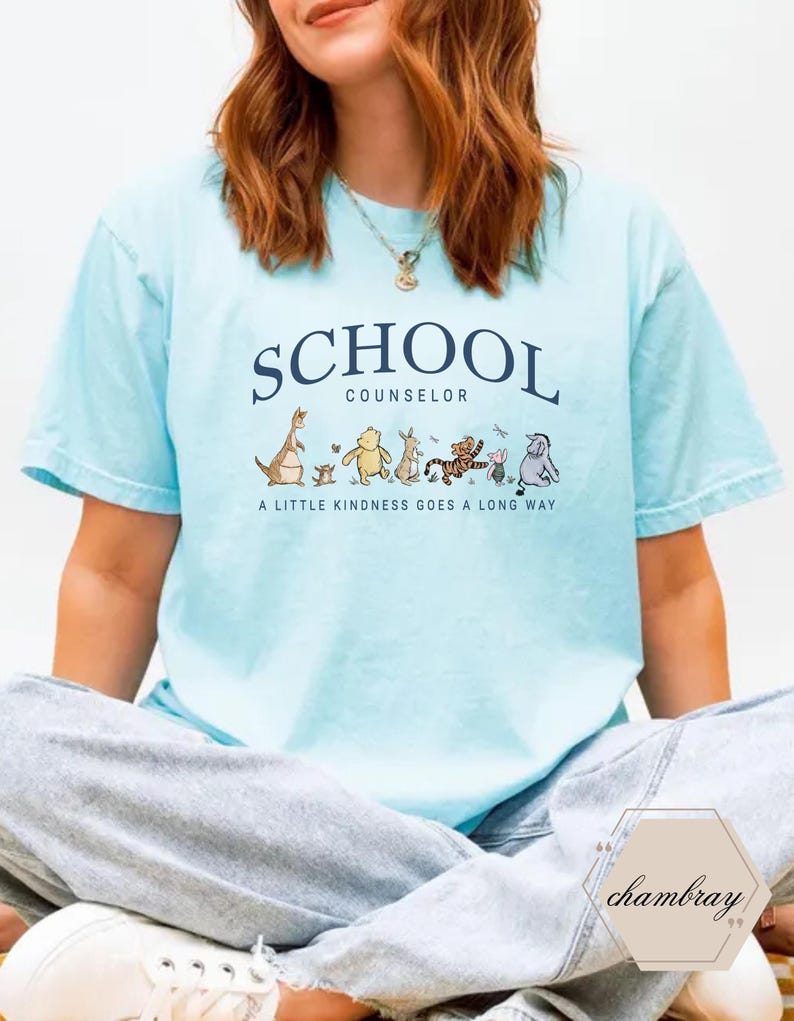 May include: A light blue short-sleeve t-shirt featuring the dark blue text SCHOOL COUNSELOR above a graphic of various cartoon animals and the text A LITTLE KINDNESS GOES A LONG WAY. The shirt has a crew neckline and a relaxed, casual fit.