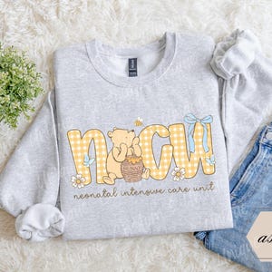 May include: A light gray sweatshirt with the letters "NICU" in yellow gingham print, featuring a cartoon bear and honey pot. The text "neonatal intensive care unit" is below the letters. A pair of blue jeans is next to the sweatshirt.