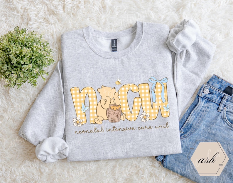 May include: A light gray sweatshirt with the letters "NICU" in yellow gingham print, featuring a cartoon bear and honey pot. The text "neonatal intensive care unit" is below the letters. A pair of blue jeans is next to the sweatshirt.
