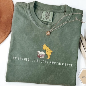 May include: A moss green t-shirt featuring a graphic of Winnie the Pooh looking at a stack of books. The text "OH BOTHER... I BOUGHT ANOTHER BOOK" is printed below the graphic. A gold necklace is draped across the shirt.