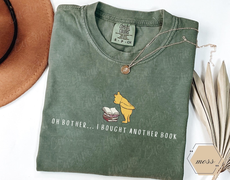 May include: A moss green t-shirt featuring a graphic of Winnie the Pooh looking at a stack of books. The text "OH BOTHER... I BOUGHT ANOTHER BOOK" is printed below the graphic. A gold necklace is draped across the shirt.
