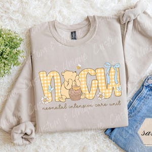 May include: A sand-colored sweatshirt with the letters "NICU" in yellow gingham print. A cartoon bear is holding a honey pot. The words "neonatal intensive care unit" are printed below the letters.
