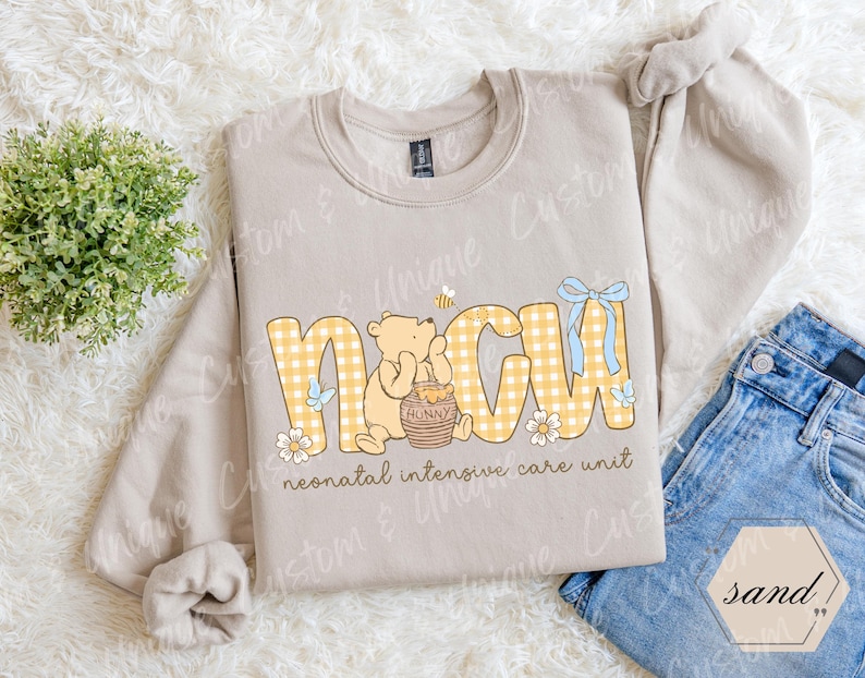 May include: A sand-colored sweatshirt with the letters "NICU" in yellow gingham print. A cartoon bear is holding a honey pot. The words "neonatal intensive care unit" are printed below the letters.