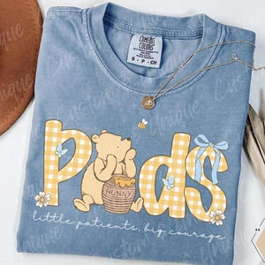 May include: A light blue t-shirt with the word "Pods" in yellow gingham letters, featuring a cartoon bear and a honey pot. The shirt also has the text "little patients, big courage". A gold necklace and a bee are also visible.