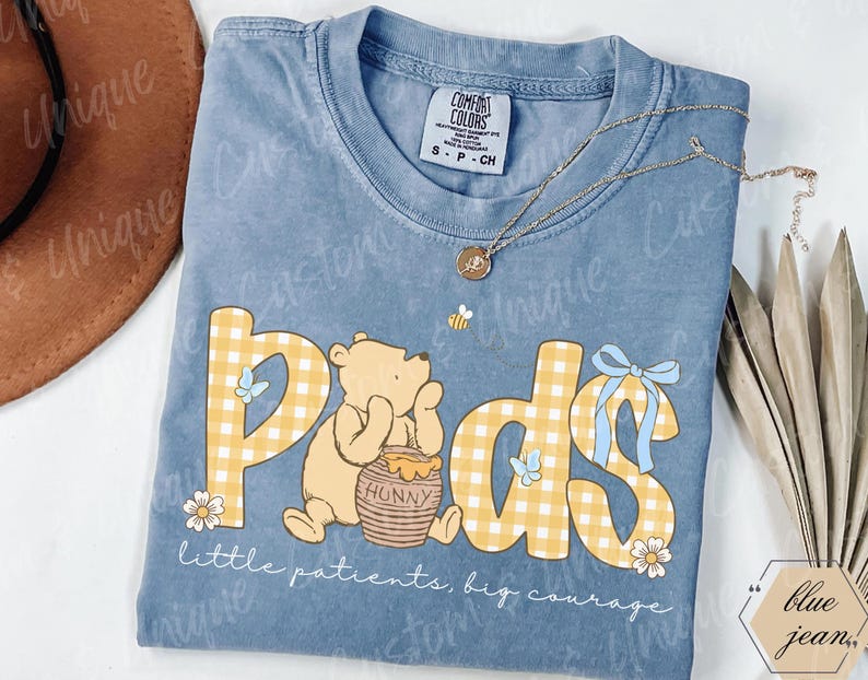 May include: A light blue t-shirt with the word "Pods" in yellow gingham letters, featuring a cartoon bear and a honey pot. The shirt also has the text "little patients, big courage". A gold necklace and a bee are also visible.