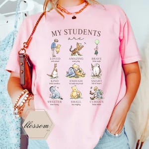 May include: Pink t-shirt with the text "MY STUDENTS are" and illustrations of Winnie the Pooh characters with positive adjectives like "LOVED" and "AMAZING". The shirt also has the word "blossom" printed on it.